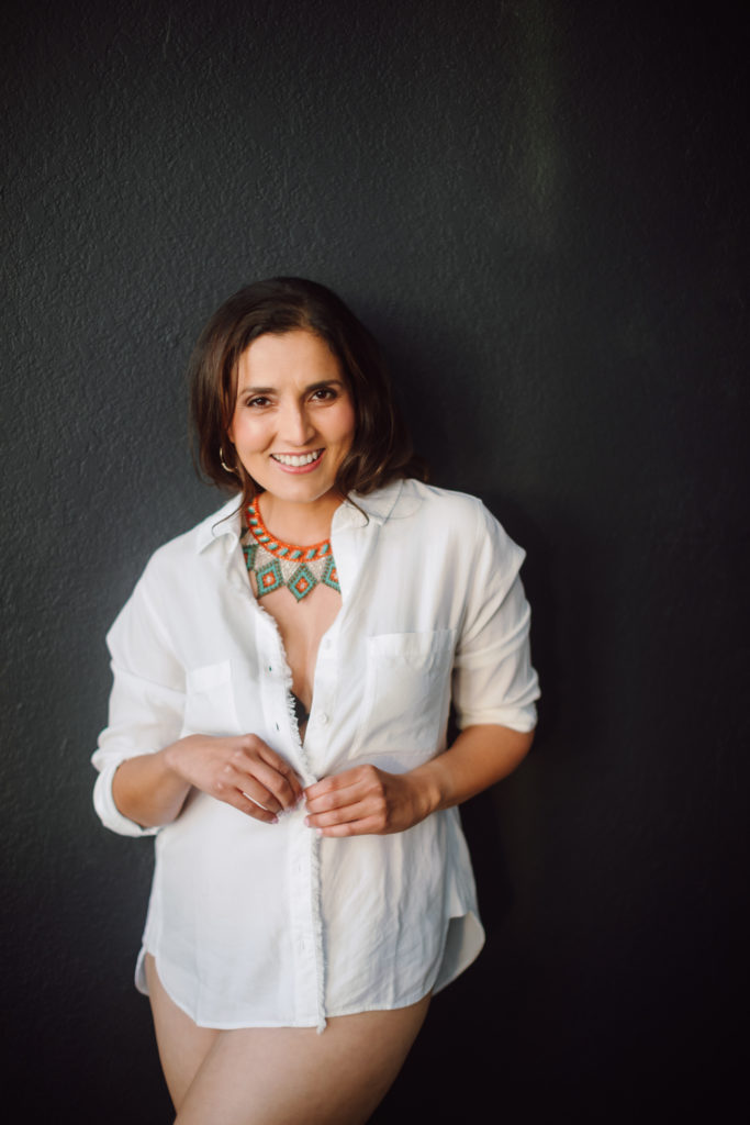 An example of what to wear for your boudoir photo shoot - a photo of a woman wearing a white button down shirt and no pants, leaning against a wall and smiling.