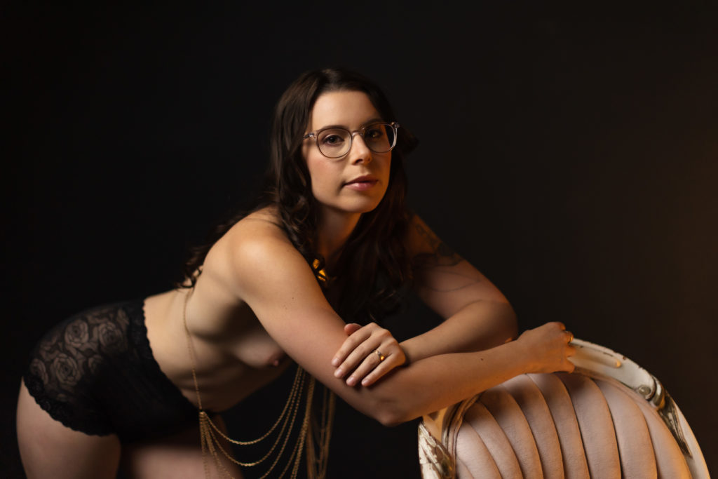 An example of what to wear to a boudoir photo session - a photo of a woman in glasses wearing a gold body chain and black underwear leaning on the back of a chair.