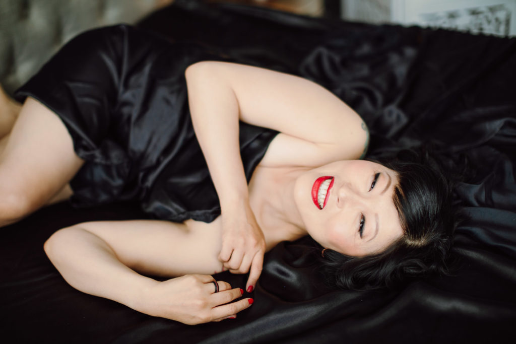 An example of what to wear to a boudoir photo shoot - a photo of a woman laying in bed, wrapped in black sheets, wearing red lipstick and smiling into the camera.