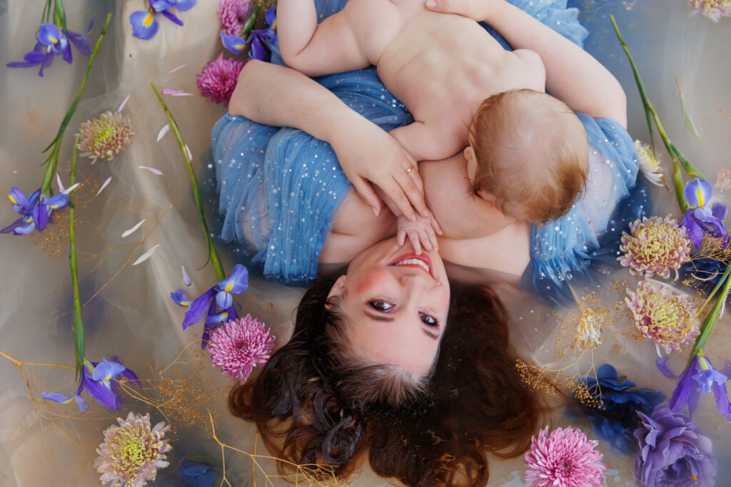 Photo of a smiling woman in a blue dress laying upside down in a flower bath. She is holding and nursing her baby. 