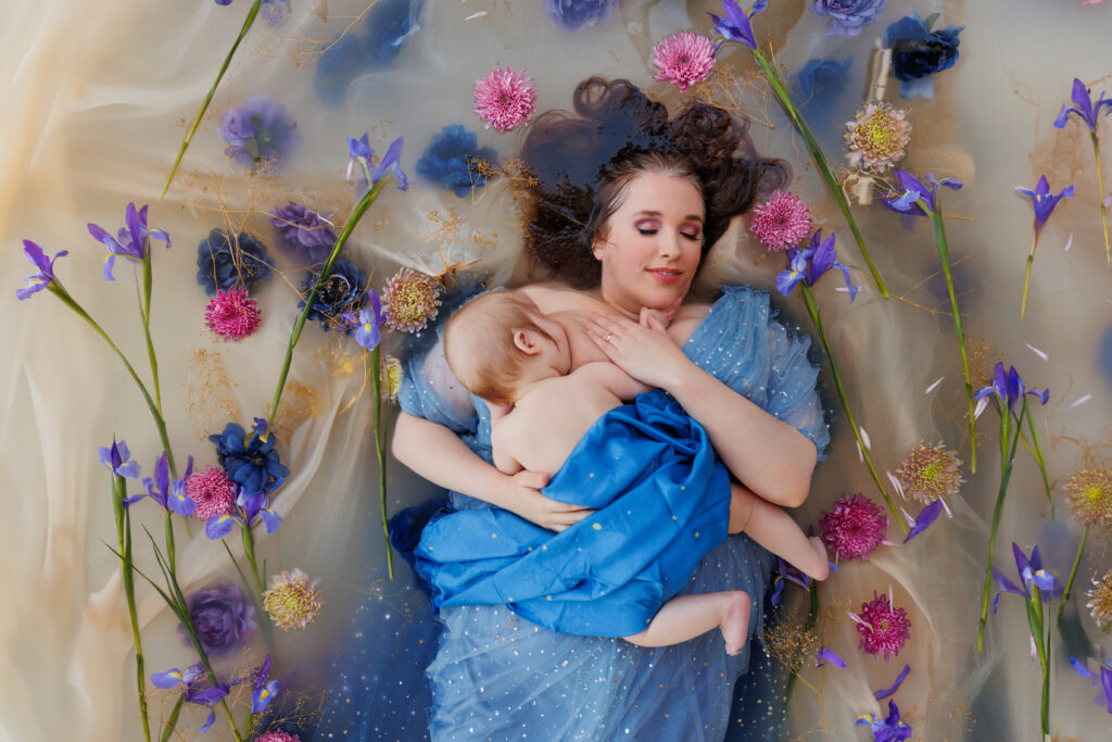 Photo of a woman with closed eyes wearing a blue dress. She is laying in a flower bath nursing her baby for a photo session. 