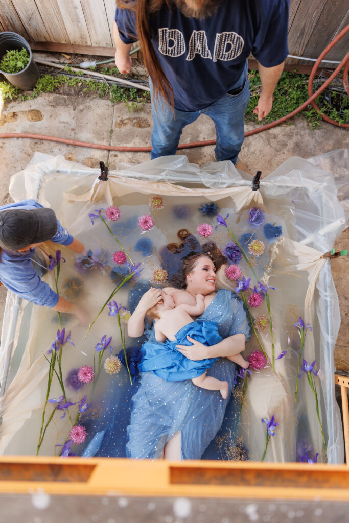 Photo of pulled-back scene of a woman nursing her baby in a bath of flowers while two other people stand next to the pool. 