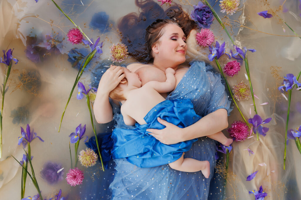 Photo of a woman with closed eyes wearing a blue dress. She is laying in a flower bath nursing her baby for a photo session. 