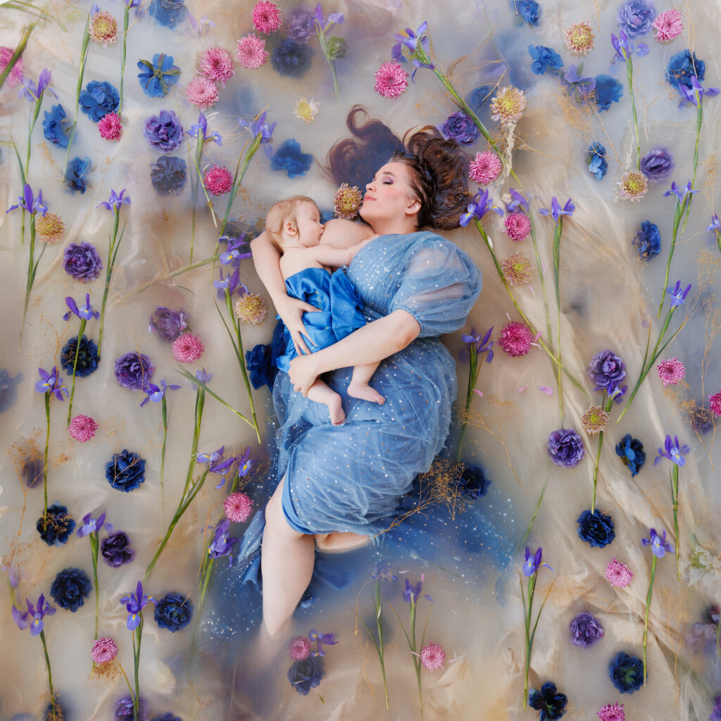 Photo from above of a woman nursing her baby in a flower bath. She is wearing blue and is surrounded by blue and purple flowers. 