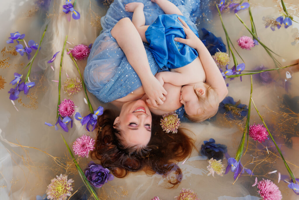Photo of a smiling woman laying upside down in a flower bath. She is holding and nursing her baby. 