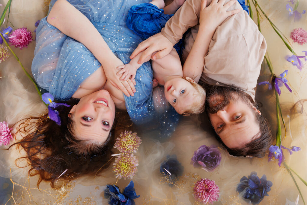 Photo of a family in a flower bath. A woman, a man, and a baby are all looking at the camera surrounded by flowers. 