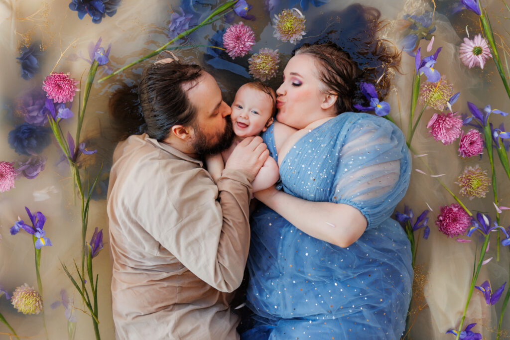 Photo of a family in a flower bath. A woman and a man are kissing their baby on the cheeks while the baby smiles at the camera. 