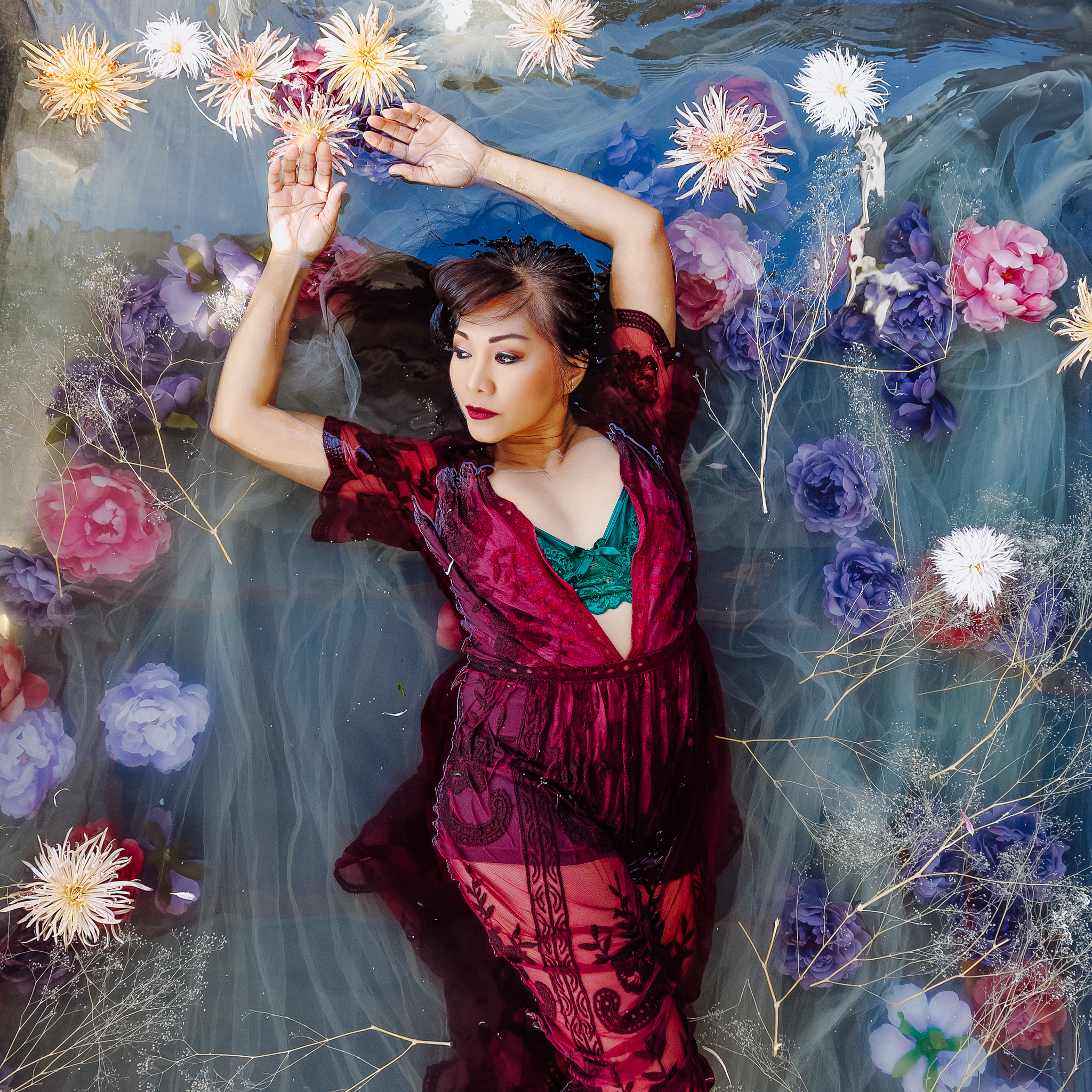 Woman laying in flower bath with her arms above her head, wearing red see-through robe and a green lacy bralette.