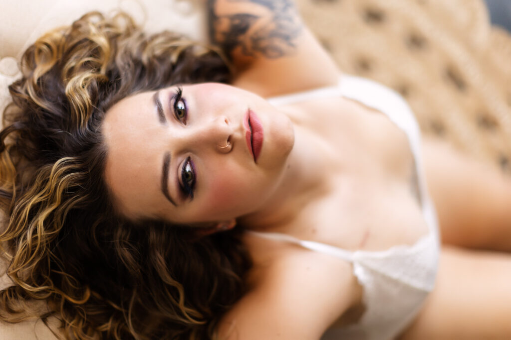 Boudoir shoot with woman with brown wavy hair in white bralette looking at camera with dark eye makeup.