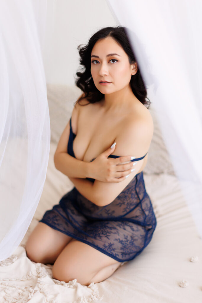 Boudoir photo of woman in black lingerie gown, sitting down in a white bed. She has her arms wrapped around her chest and is looking at the camera.